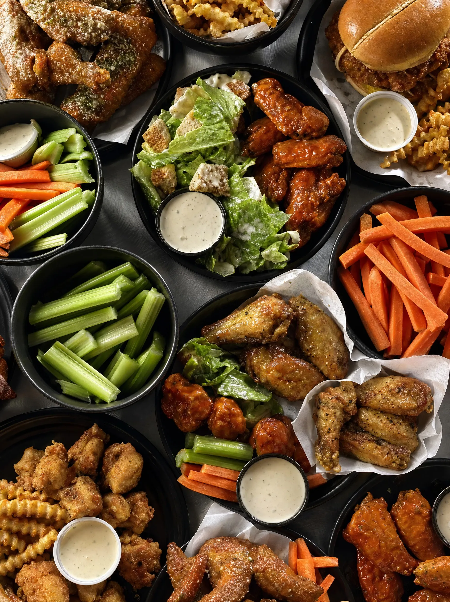 A Wing Kings group order laid out across a table: buffalo wings, dry-rub wings, boneless bites, crinkle fries, chicken sandwiches, caesar salads, carrot and celery sticks and ranch cups